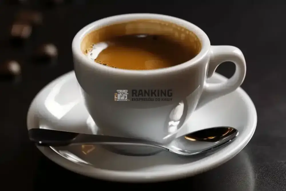 Biała filiżanka z espresso na białym spodku, obok której leży mała łyżeczka. Całość ustawiona na ciemnym tle, z delikatnymi refleksami światła na powierzchni filiżanki i spodka.